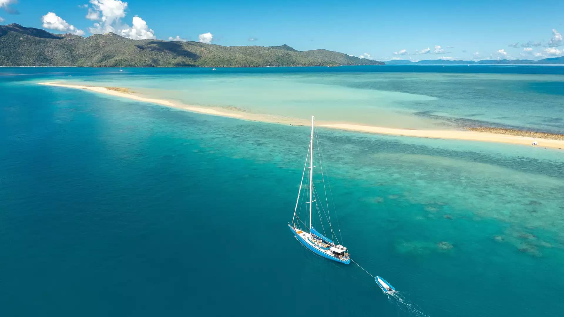 A high-angle aerial view of a large blue sailboat anchored in crystal-clear water next to the long, narrow white sandspit of Langford Island in the Whitsundays.