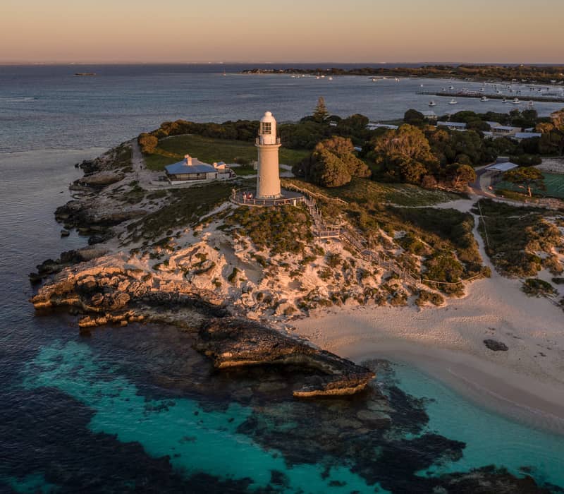 A high-angle aerial shot showing a tall white lighthouse standing on a grassy, rocky headland. In the foreground, the dark rocks meet clear turquoise water, while in the background, a blue bay is filled with white boats at anchor.