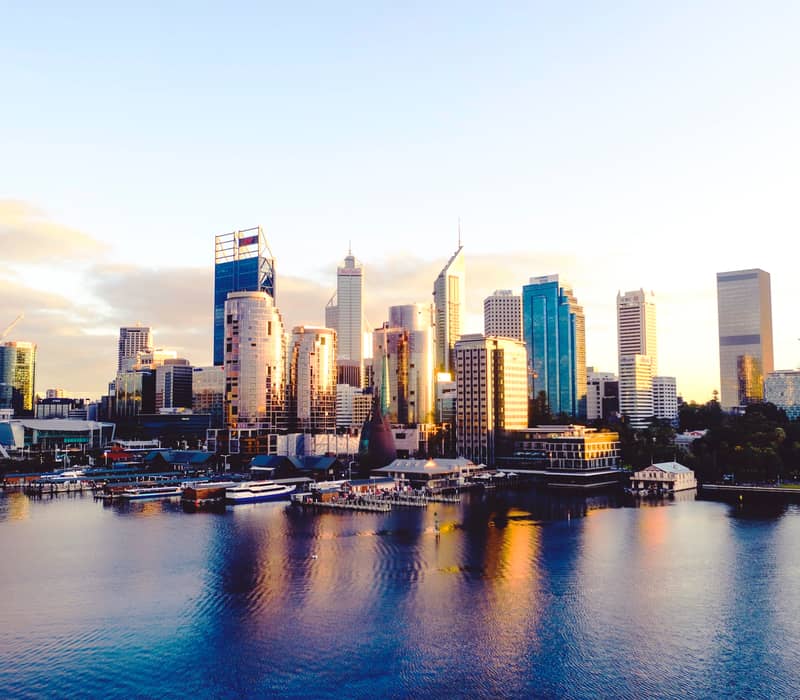 A wide landscape shot taken at sunrise, showing the tall, glass skyscrapers of the Perth CBD reflecting golden light. In the foreground, the calm blue water of Elizabeth Quay features a modern pedestrian bridge and several ferries docked at the pier.