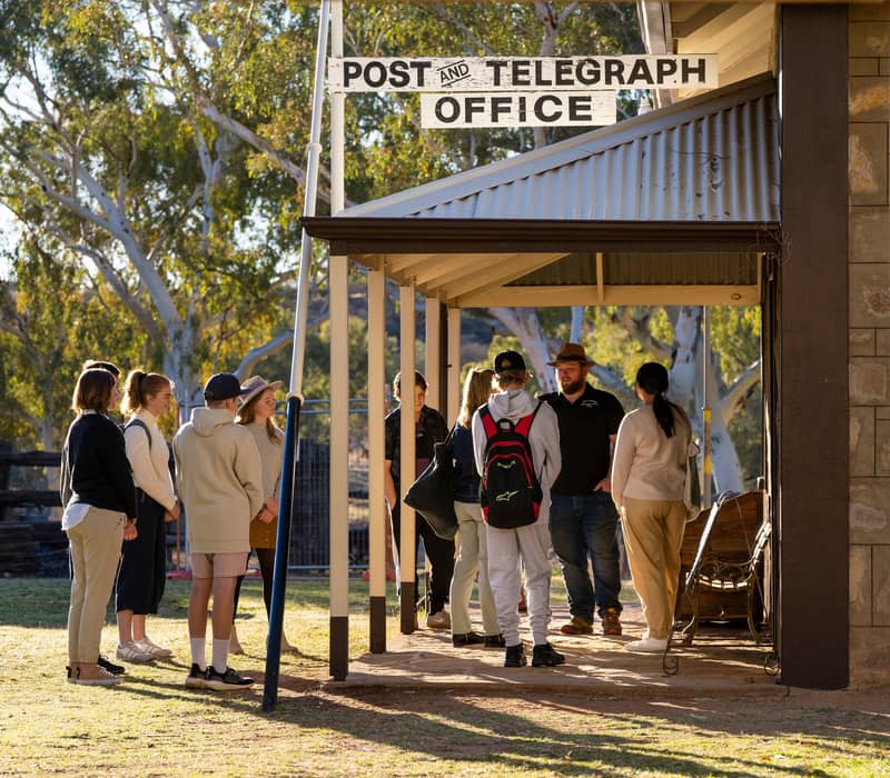A tour guide in a hat speaking to a group of young travelers outside a historic stone post office building.