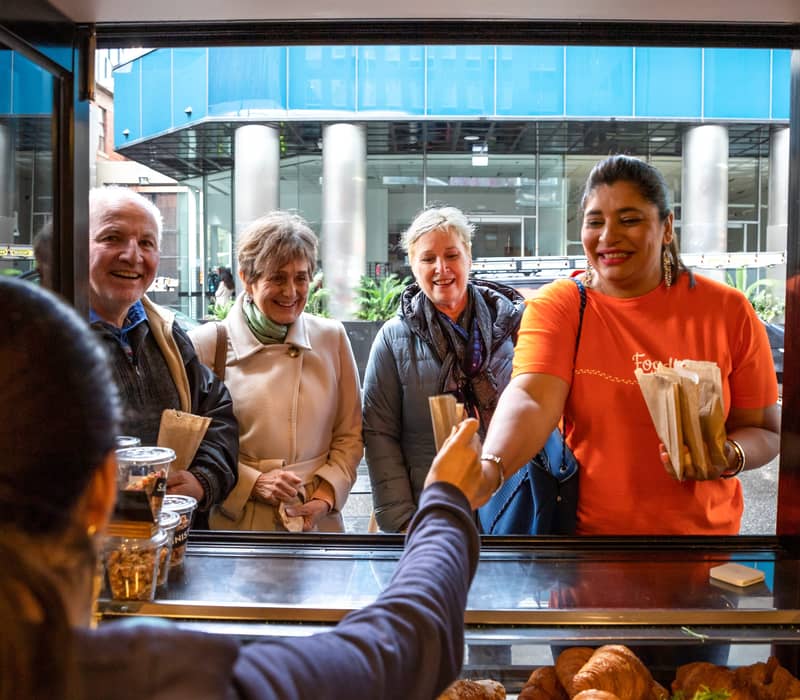 A smiling tour guide in an orange shirt hands a paper bag to a customer through a café service window in a Melbourne lane.