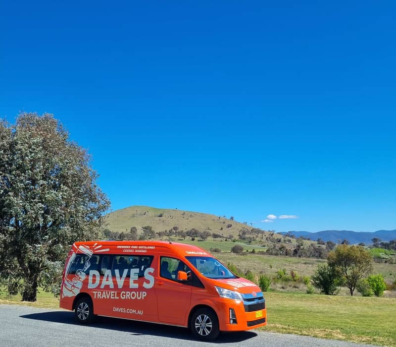 An orange passenger van parked on a roadside with rolling green hills and a blue sky in the background.