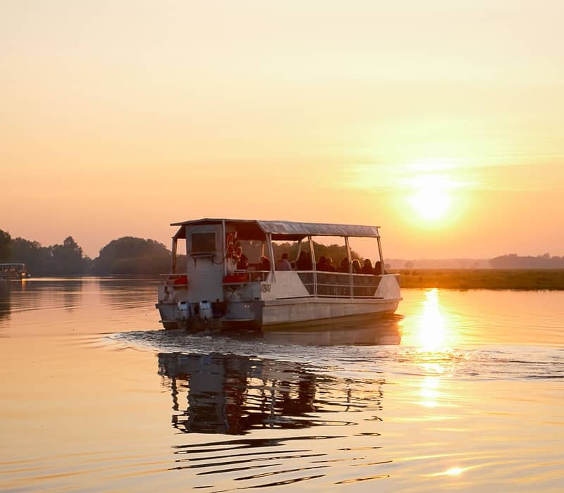 A tour boat filled with people silhouetted against a bright golden sunset on a calm river.