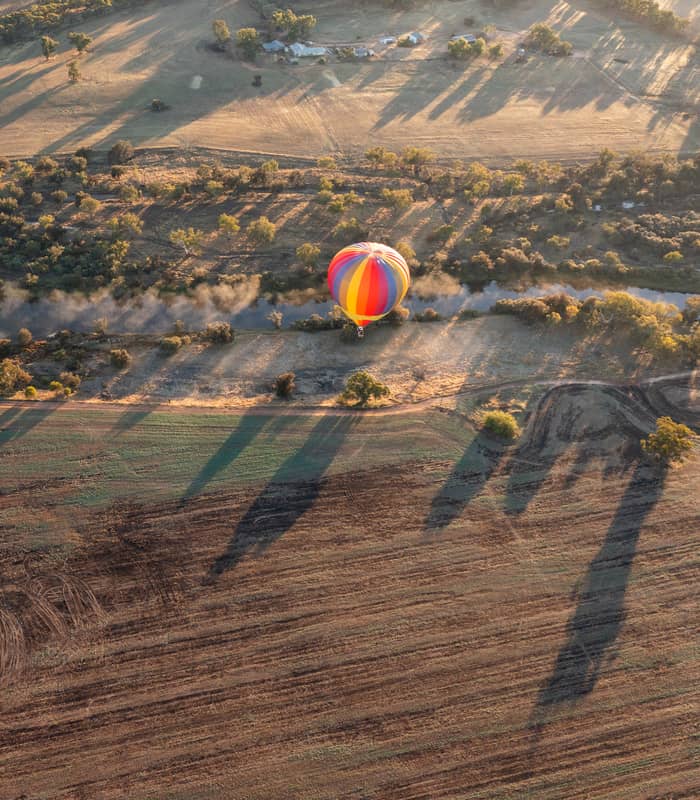 A high-angle aerial shot showing a large, colorful striped hot air balloon (red, yellow, blue, and orange) floating over a calm river and rural landscape. Long shadows from the morning sun stretch across ploughed fields and green trees, with a light morning mist rising from the water.