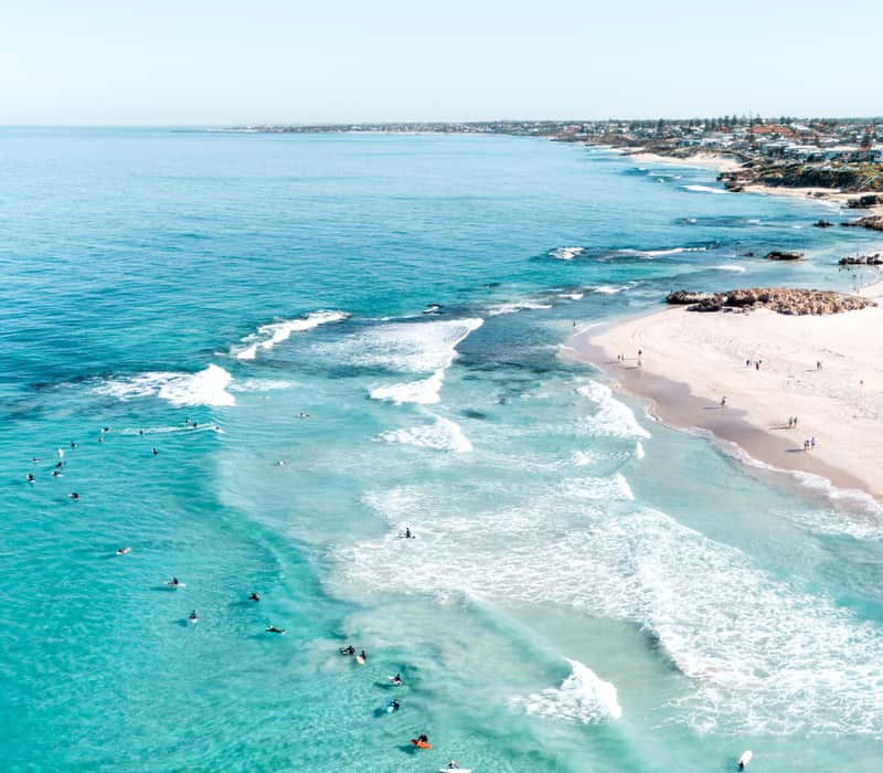 A vertical aerial photograph of a pale blue ocean with small white waves rolling toward a white sand beach. Dozens of surfers on boards are scattered throughout the water, and several people are walking along the shoreline.