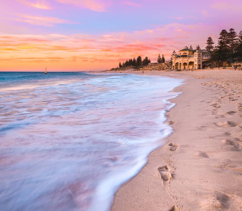 A vertical beach photograph at sunset with a pink and orange sky. Footprints lead along the wet sand in the foreground toward the Indiana Teahouse, a grand yellow building on the shore. Soft white waves are rolling onto the beach under the colorful evenin