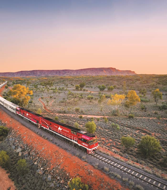 High-angle view of the long Ghan train with its signature red engines curving through a vast desert landscape toward distant mountains.