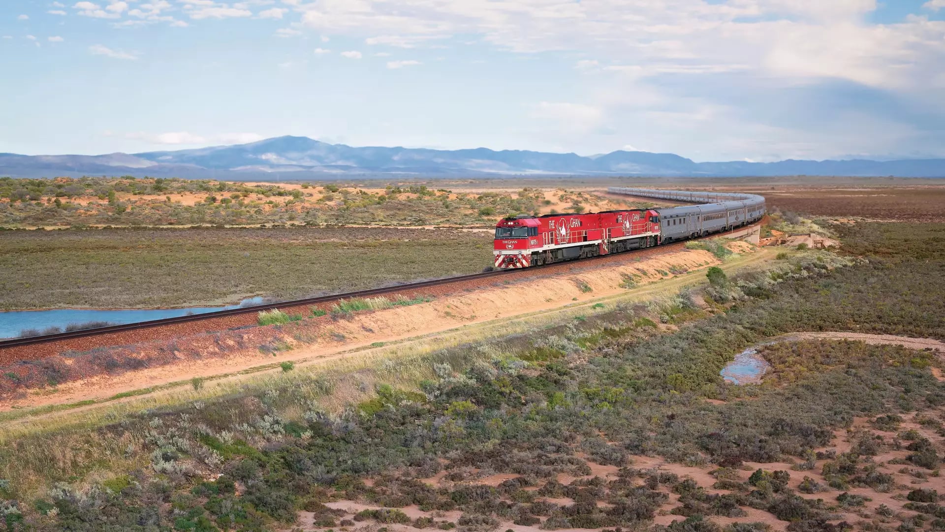 High-angle view of the long Ghan train with its signature red engines curving through a vast desert landscape toward distant mountains.