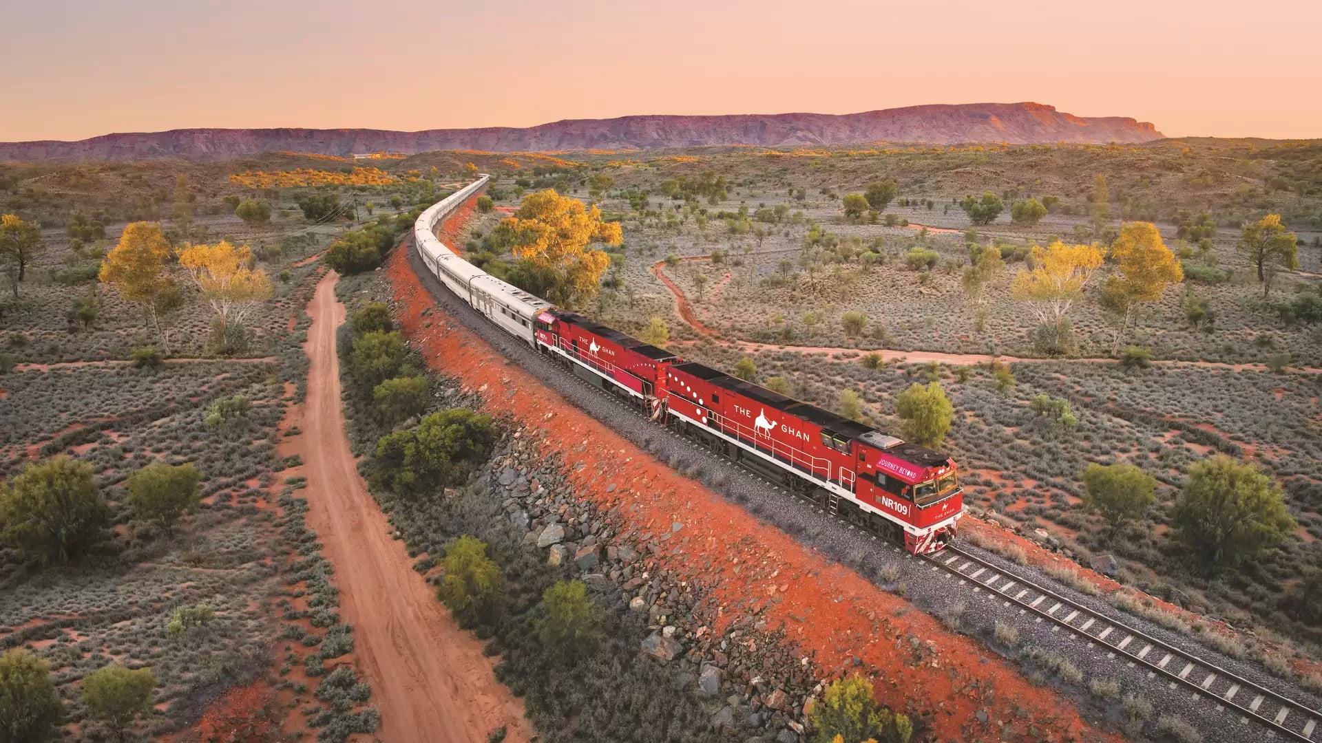 High-angle view of the long Ghan train with its signature red engines curving through a vast desert landscape toward distant mountains.