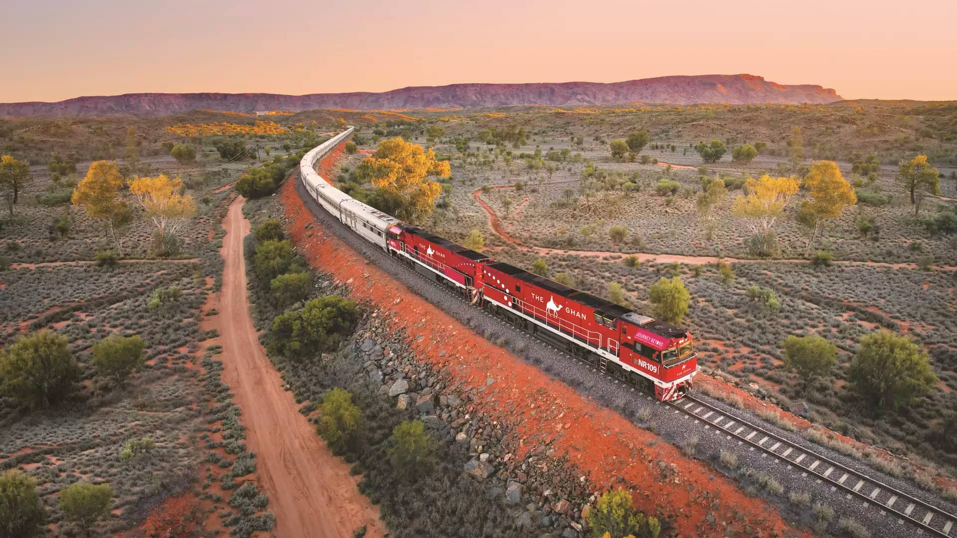 High-angle view of the long Ghan train with its signature red engines curving through a vast desert landscape toward distant mountains.