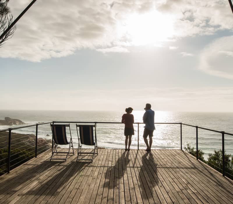 Silhouette of a couple standing on a wooden balcony overlooking the ocean with bright sunlight reflecting off the water.