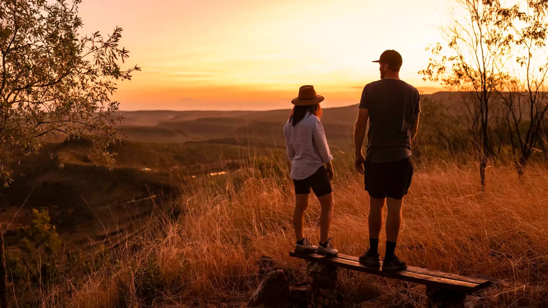 A man and a woman stand on a wooden bench on a grassy hill, looking out at a vast, dry landscape of rolling hills and ranges during a golden-hour sunset