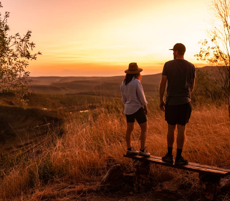 A man and a woman stand on a wooden bench on a grassy hill, looking out at a vast, dry landscape of rolling hills and ranges during a golden-hour sunset