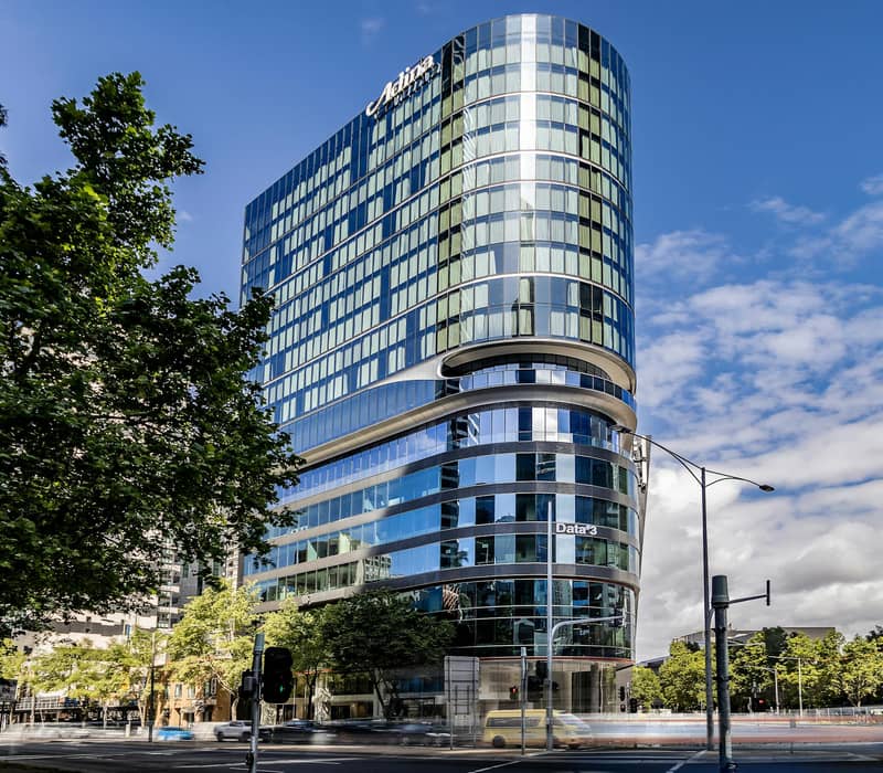 Tall modern glass hotel building with trees and street in foreground under blue sky