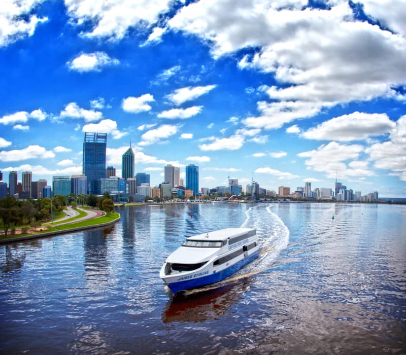 High-angle view of a white ferry on a wide river with a city skyline and bright blue sky with fluffy white clouds.