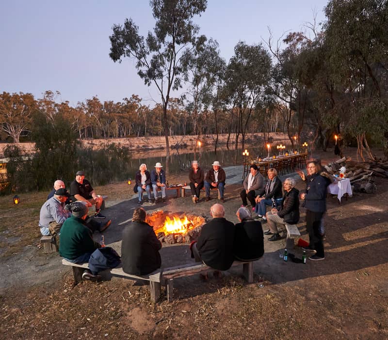 Guests gathered around a central campfire at dusk for a unique dining experience on the banks of the Murray River at Echuca Moama.