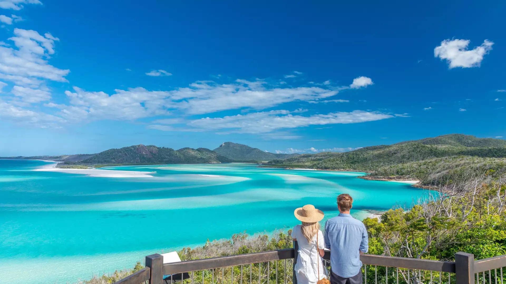A man and woman looking out from a wooden balcony over the turquoise swirling sands of Hill Inlet.