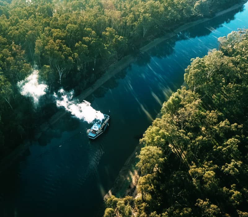 High-angle drone photograph of the paddlesteamer PS Emmylou cruising a straight stretch of the Murray River surrounded by forest.