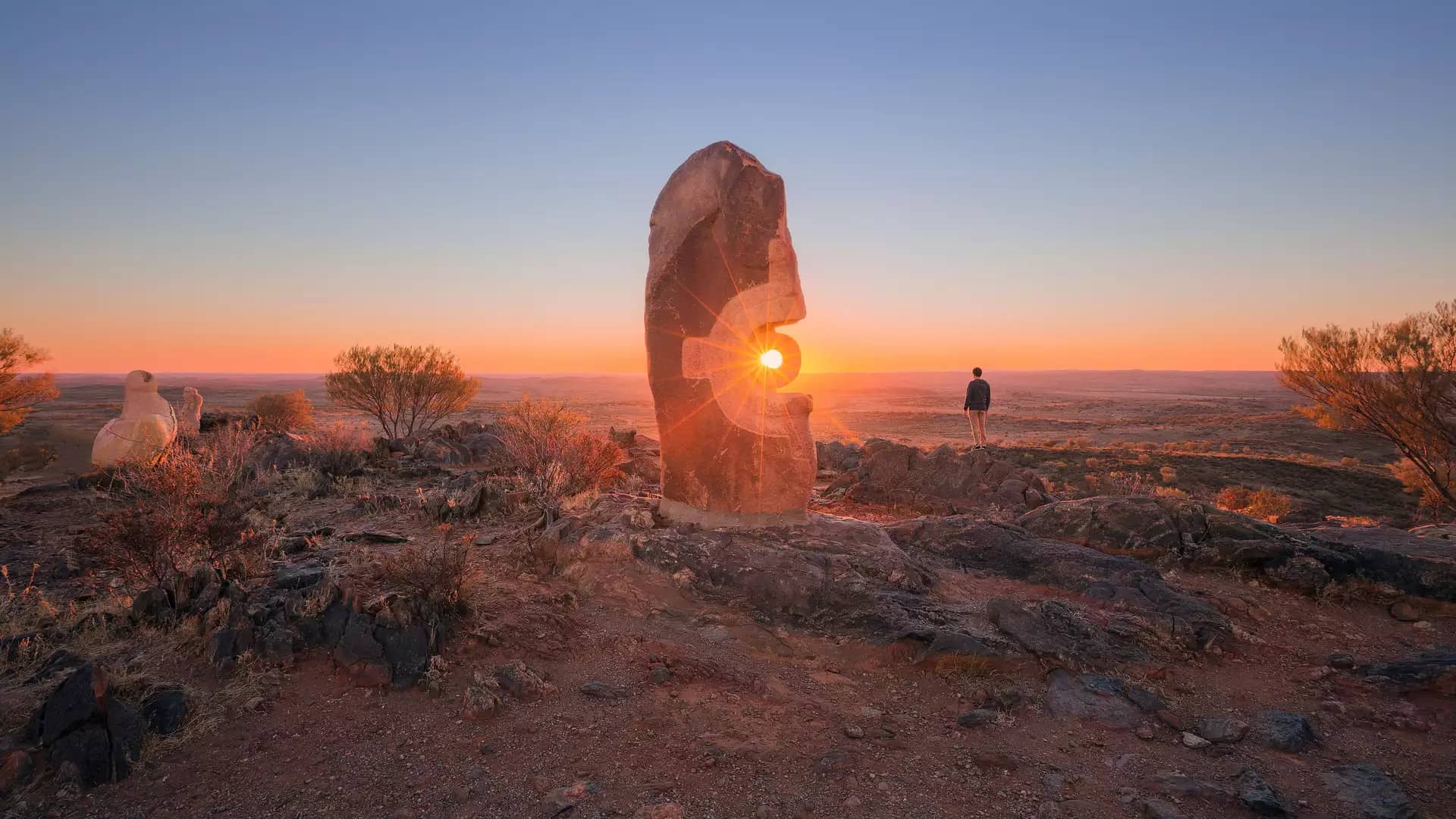 Carved sandstone sculptures standing on a hilltop overlooking the desert plains of Broken Hill under a clear sky.