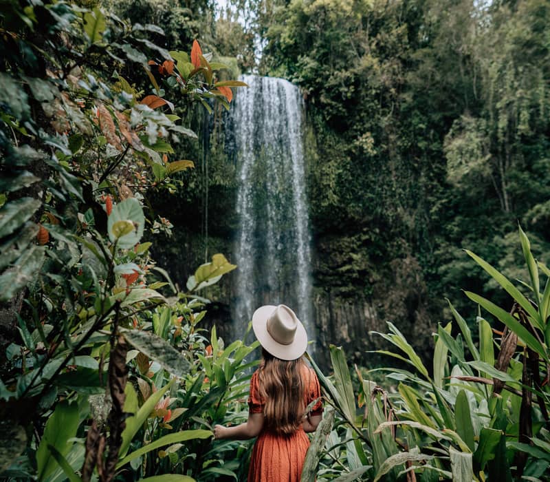A woman in an orange dress and hat stands before the powerful drop of Millaa Millaa Falls.