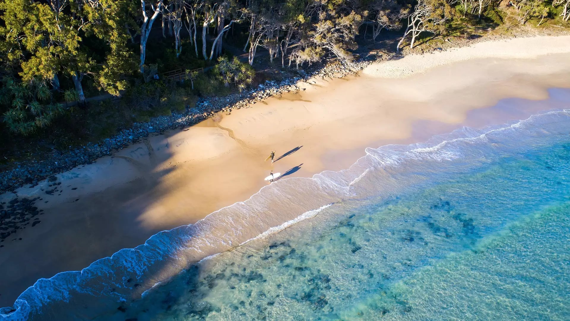 A top-down aerial view of two surfers walking across the sand at Tea Tree Bay, highlighting the crystal-clear water and rocky shoreline.