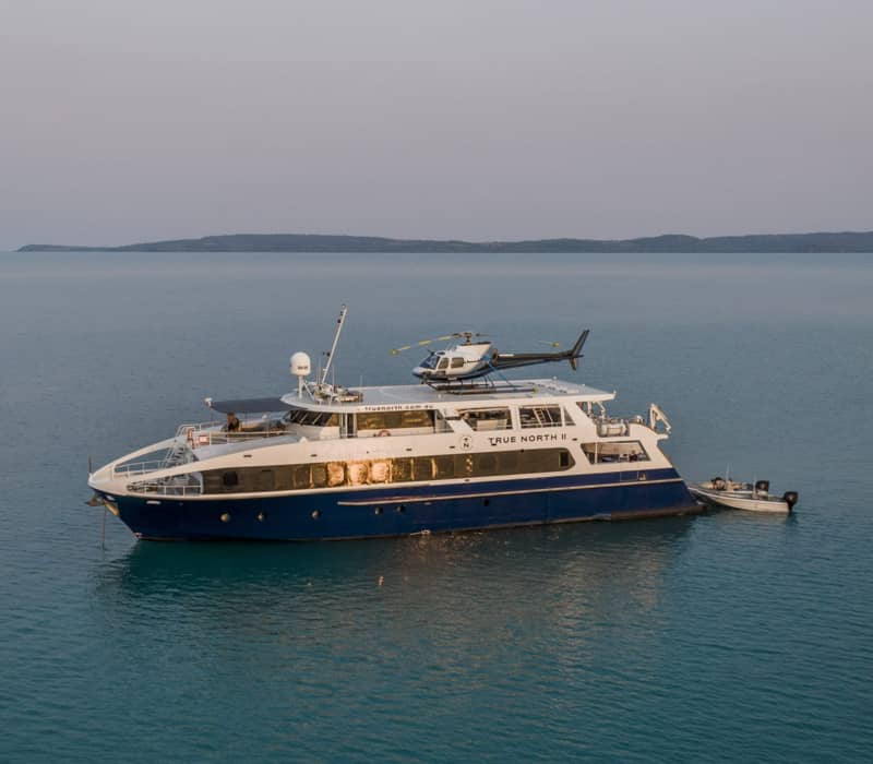The blue and white True North II cruise ship at sunset with a helicopter on deck and a tender boat attached to the rear.