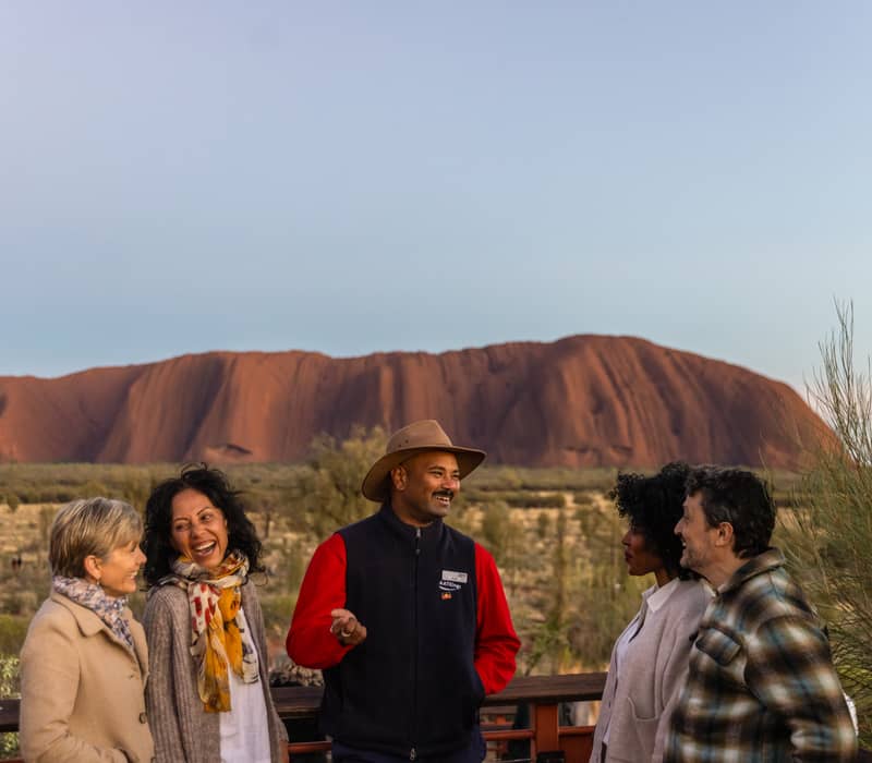 A group of people laughing together at a lookout with the massive red rock of Uluru in the background.