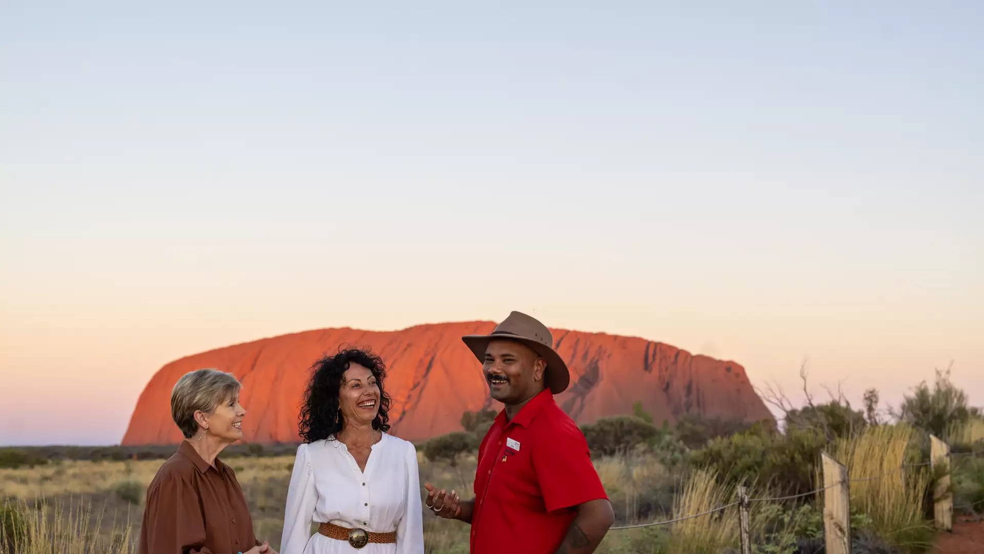 A guide talking to two women with the vibrant red rock of Uluru in the background at sunset.