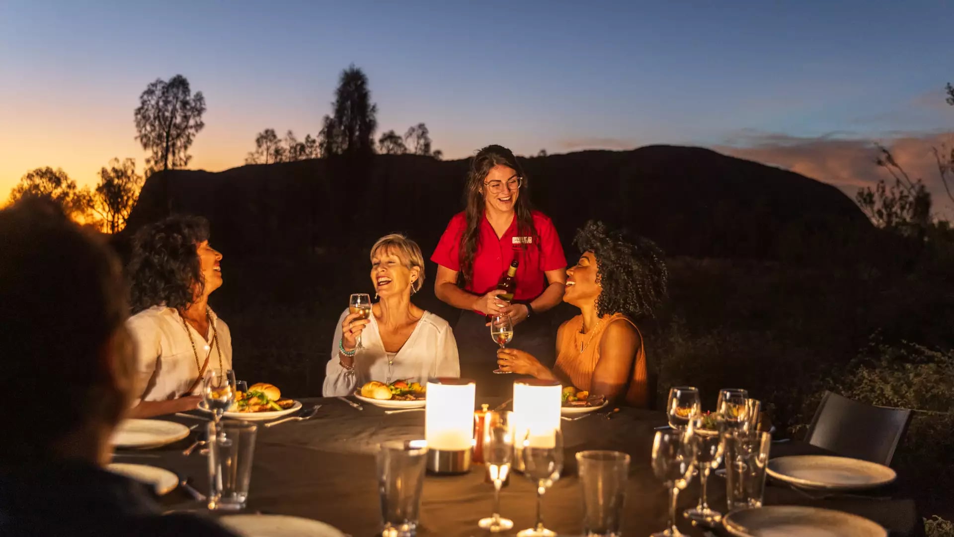 Travellers enjoying a candlelit dinner and wine at an outdoor table in the Red Centre at dusk.