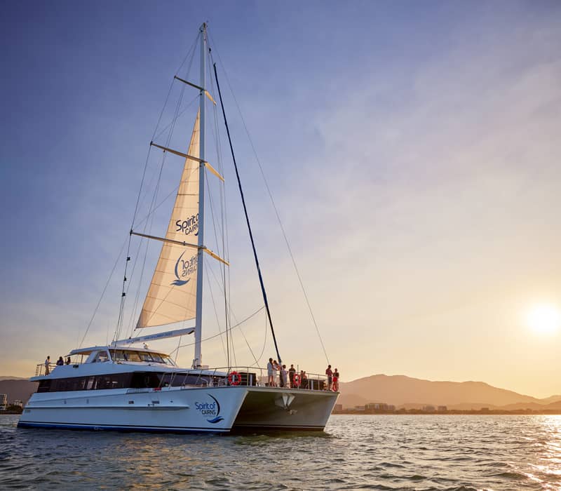 A large white sailing catamaran with passengers on deck at sunset with the Cairns mountains in the background.