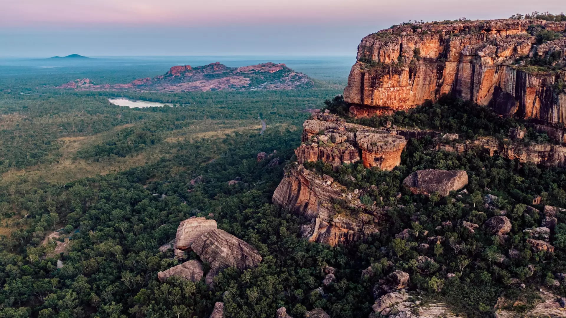 A breathtaking aerial shot of the rugged red sandstone cliffs of Burrungkuy (Nourlangie) at dusk, with the vast, green savanna woodlands of Kakadu National Park stretching toward the horizon under a soft pink and purple sky.