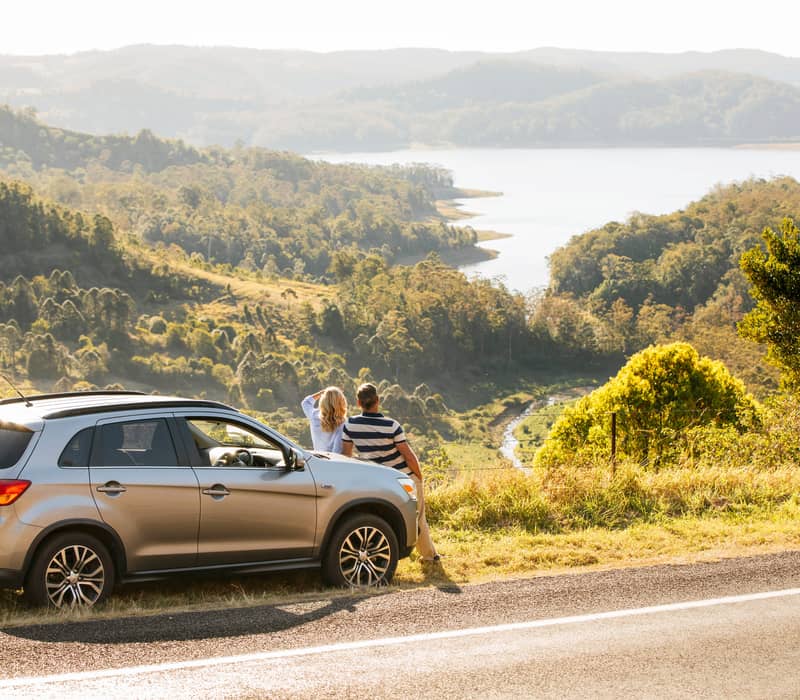 A silver SUV parked on the side of a mountain road, with a man and woman leaning against it looking out over a blue lake in a green valley.