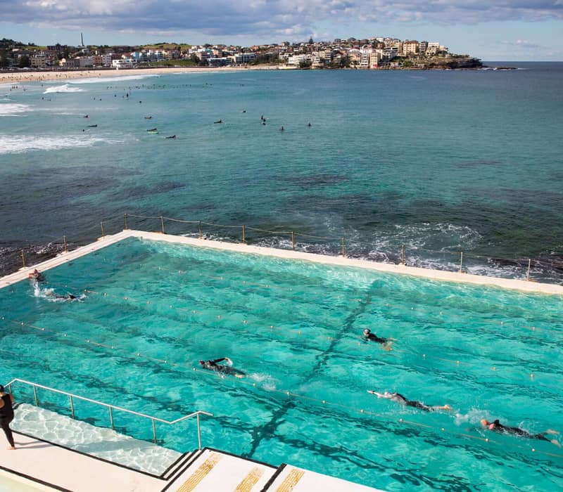 High-angle view of people swimming laps in a bright turquoise rectangular ocean pool built into rocky cliffs next to the sea at Bondi Beach.