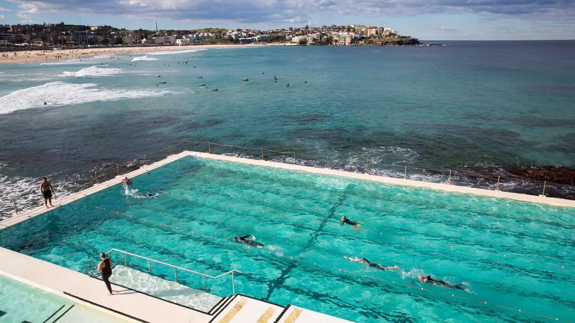 High-angle view of people swimming laps in a bright turquoise rectangular ocean pool built into rocky cliffs next to the sea at Bondi Beach.
