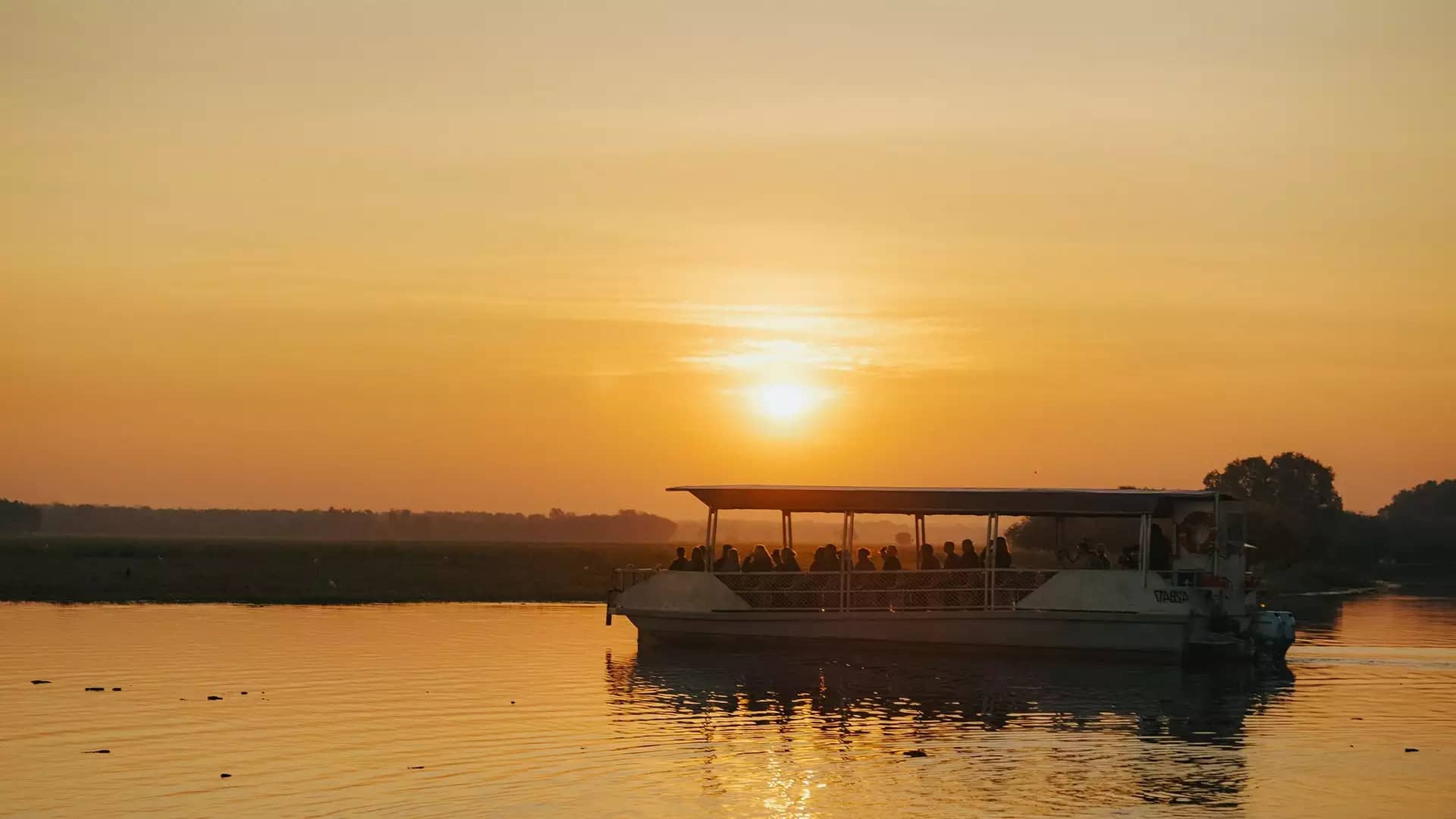 A passenger cruise boat glides across the tranquil Yellow Water Billabong in Kakadu National Park during a vibrant sunset.