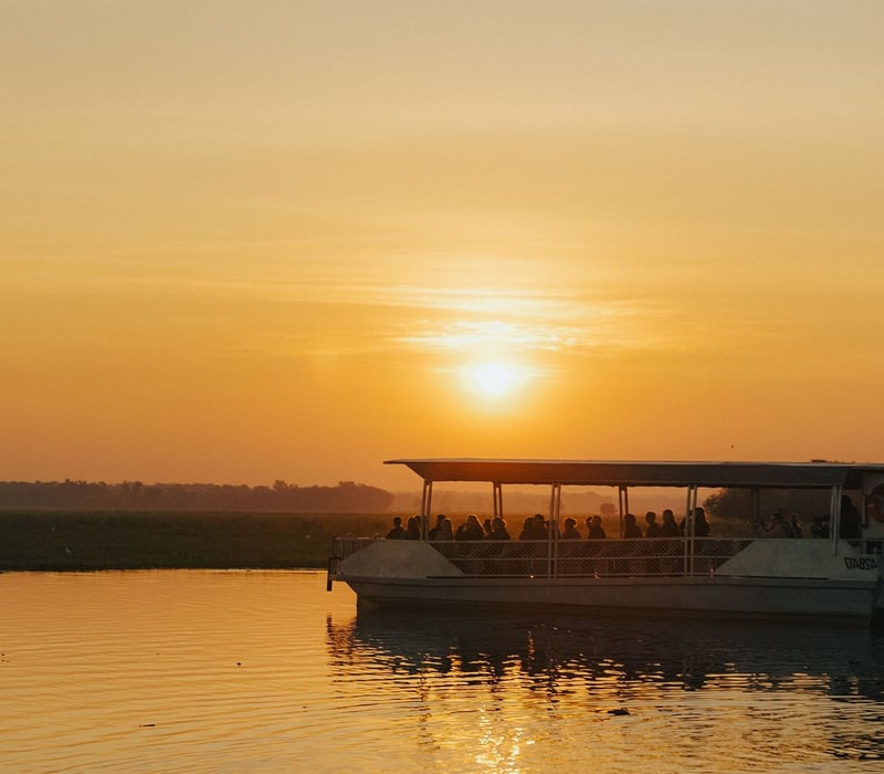 A passenger cruise boat glides across the tranquil Yellow Water Billabong in Kakadu National Park during a vibrant sunset.