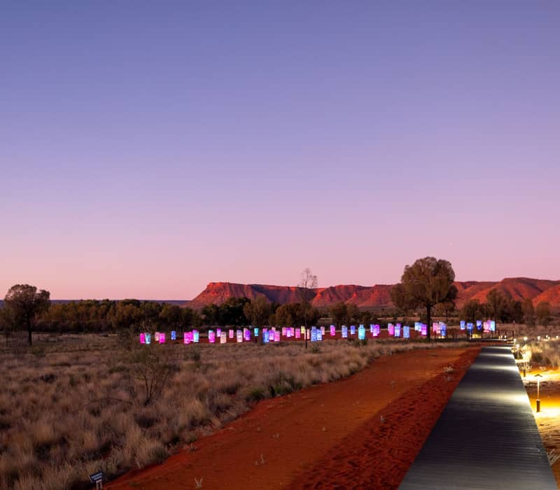 A wooden boardwalk with small floor lights leads through red sand toward a field of glowing, colorful glass towers in the desert at dusk.