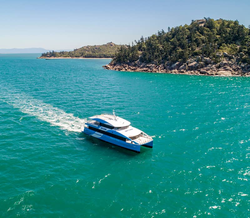 A large blue and white SeaLink passenger ferry cruising through calm turquoise water near the Magnetic Island coastline.