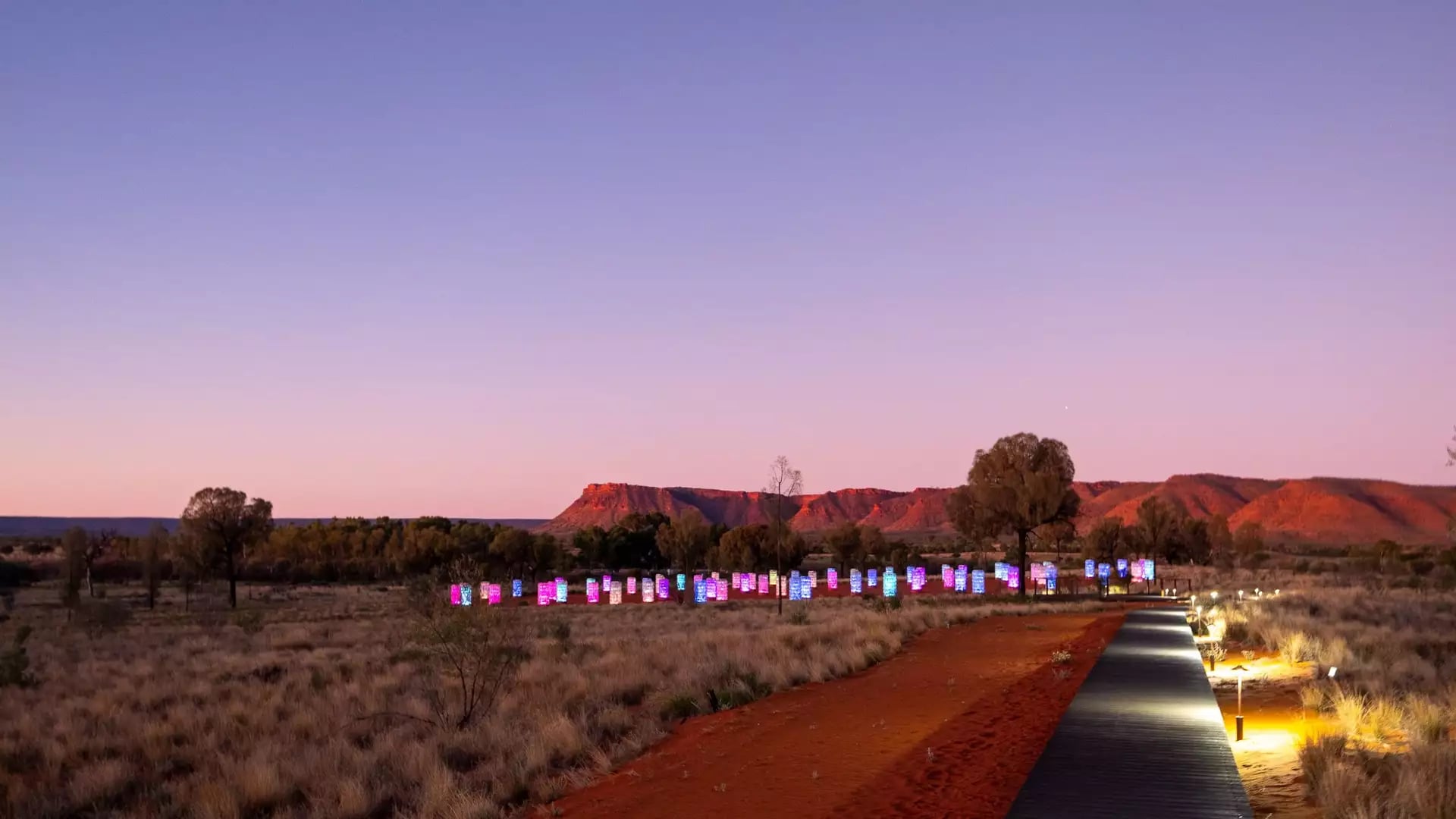 A wooden boardwalk with small floor lights leads through red sand toward a field of glowing, colorful glass towers in the desert at dusk.