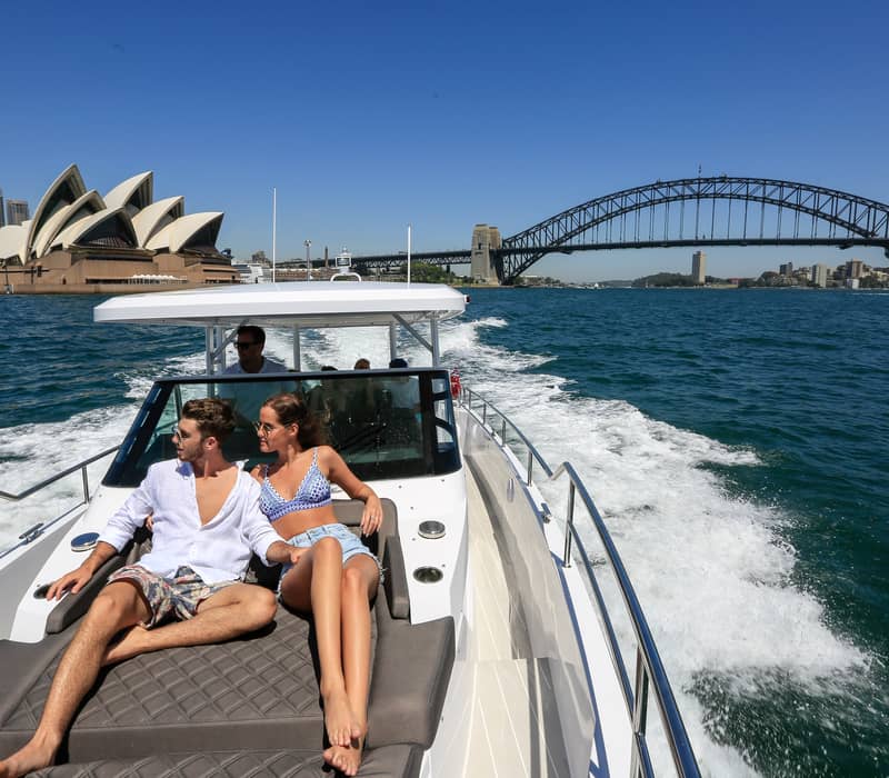Couple sitting on a boat deck cruising past the Sydney Opera House and Harbour Bridge.