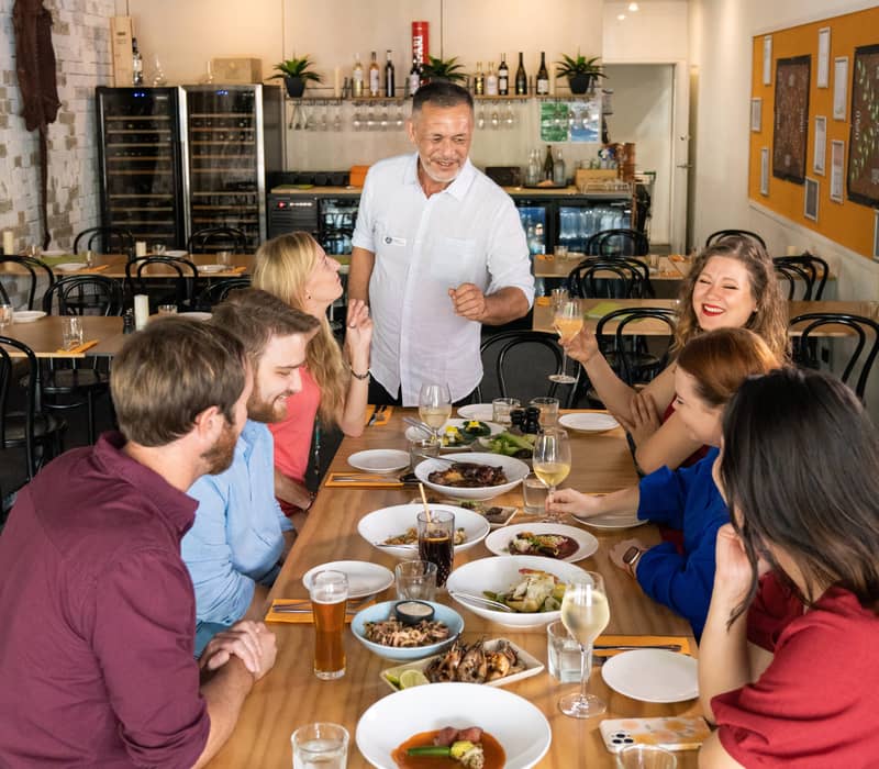 A group of smiling people sit around a long wooden table covered in various dishes and wine glasses, interacting with a male server in a white shirt.