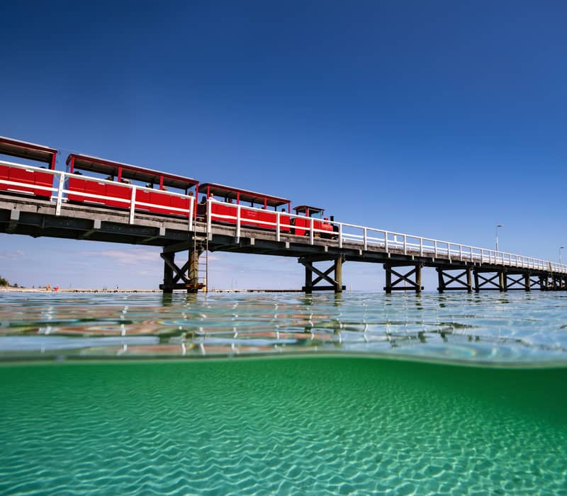 A split-level photograph shows the red Busselton Jetty tourist train on the tracks above and the sun-dappled, clear green water of Geographe Bay below in Western Australia.