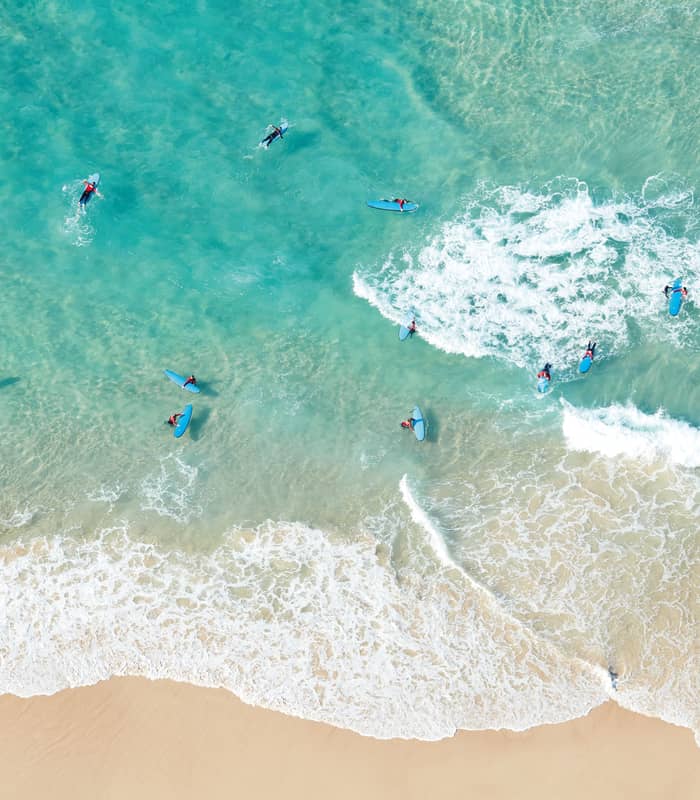 A top-down aerial view of several people on blue surfboards scattered in clear turquoise water with white foam from breaking waves.