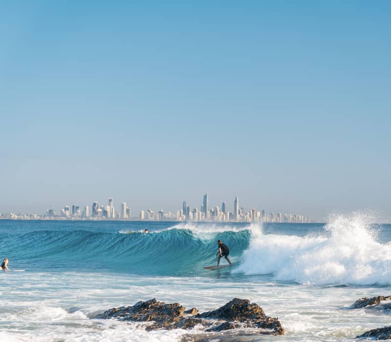 A surfer skillfully navigates a peeling blue wave at the famous Currumbin Alley, framed by the rugged rocks of the foreground and the distant city skyline.