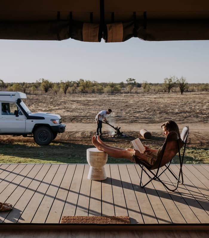 A woman reads a book on a timber deck of a luxury safari-style tent overlooking the vast Longreach plains at sunset.