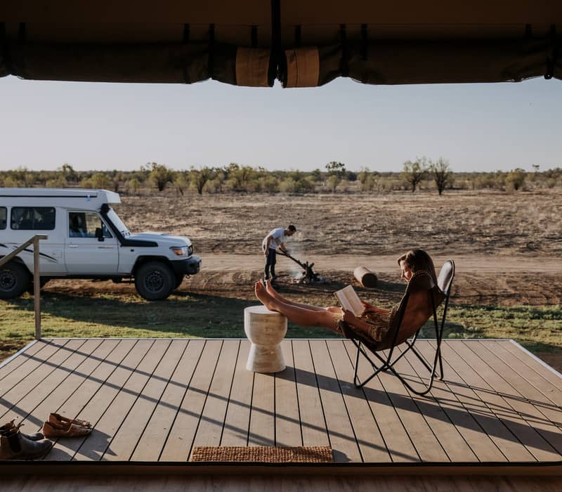 A woman reads a book on a timber deck of a luxury safari-style tent overlooking the vast Longreach plains at sunset.