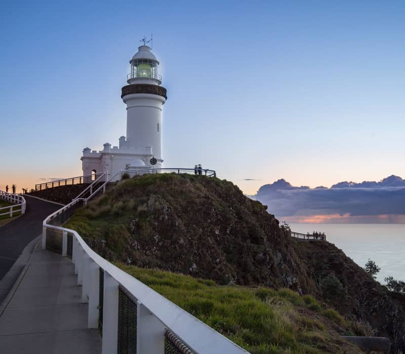 A white lighthouse on a grassy hill with a paved walking path along a cliff edge under a clear dawn sky.