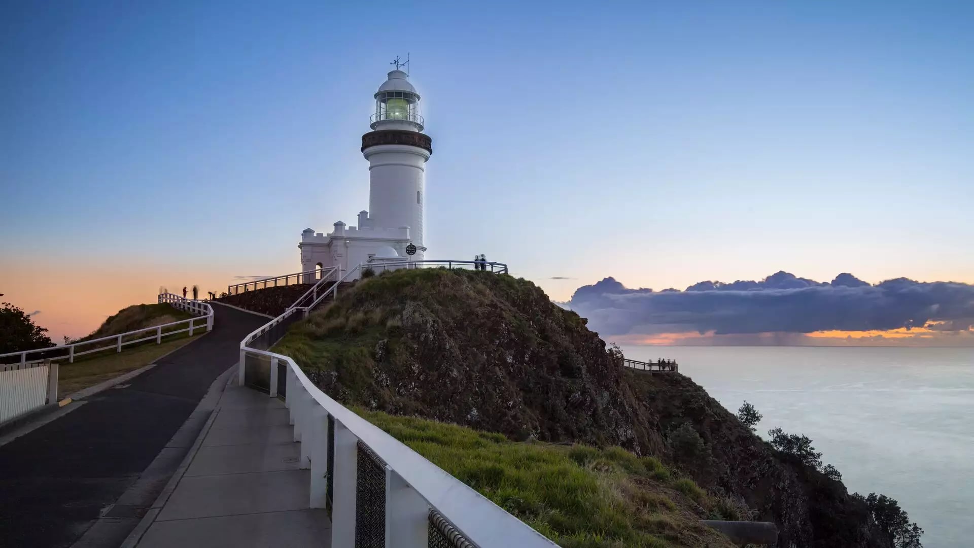 A white lighthouse on a grassy hill with a paved walking path along a cliff edge under a clear dawn sky.