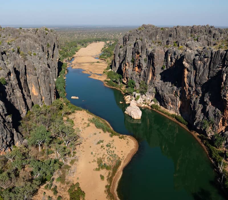 High-angle drone shot of a green river snaking through a deep canyon with dark grey vertical rock walls and green trees.