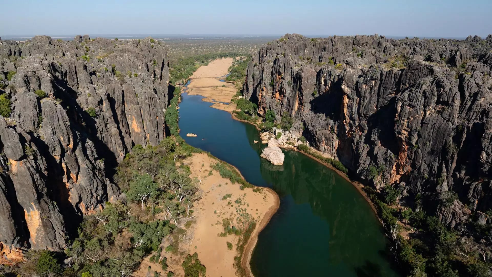 High-angle drone shot of a green river snaking through a deep canyon with dark grey vertical rock walls and green trees.